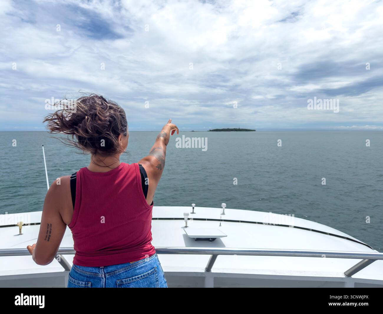Una donna sta su una barca a Cairns, in Australia, puntando verso un'isola lontana sotto un cielo parzialmente nuvoloso. I suoi capelli vengono soffiati dal vento, catturando un Foto Stock