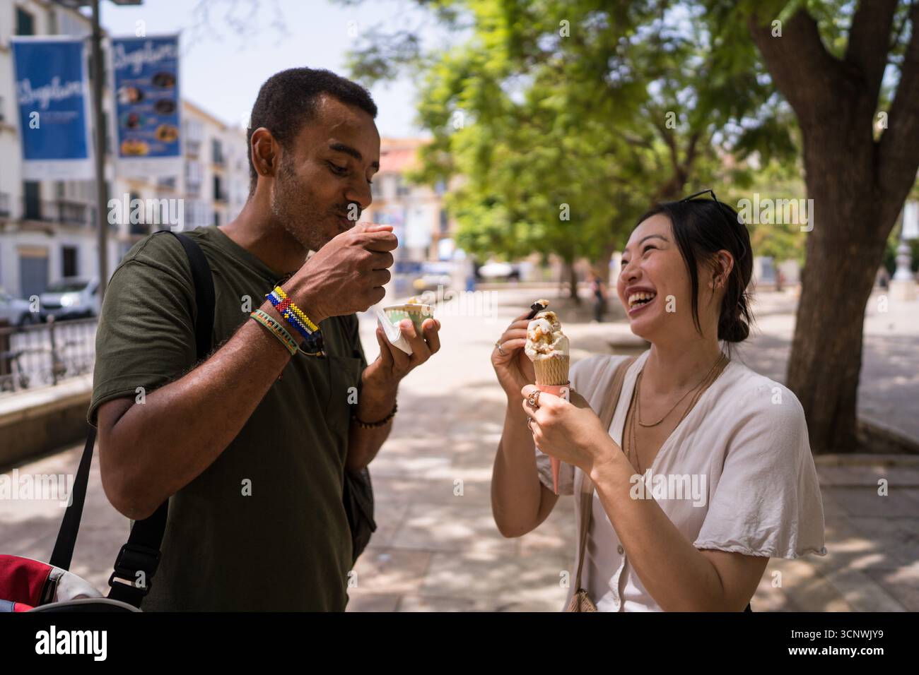 Una coppia gioiosa condivide un momento delizioso gustando i coni di gelato in una vivace piazza della città durante una giornata di sole, catturando l'essenza del divertimento estivo a. Foto Stock