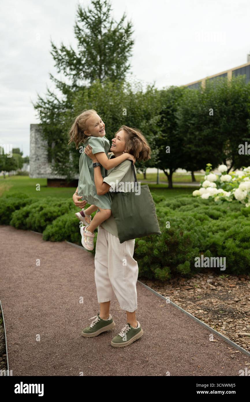 Una madre gioiosa solleva la figlia ridendo in aria mentre giocano in un bellissimo parco giardino. Circondati dal verde, il loro momento cattura il puro caos Foto Stock