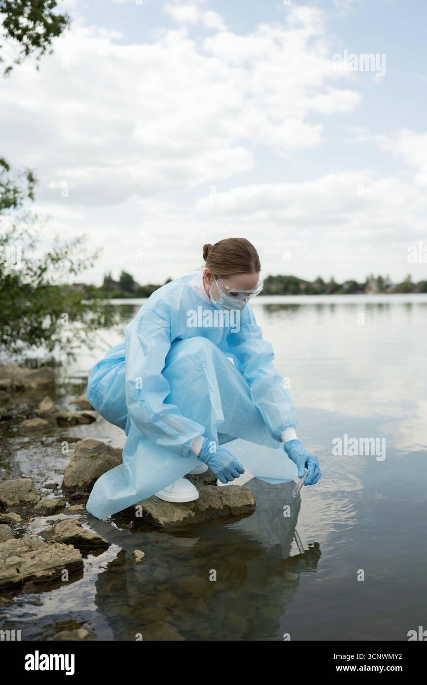 Una giovane biologa in attrezzatura protettiva conduce ricerche ecologiche in un lago sereno, raccogliendo campioni d'acqua per studiare gli impatti ambientali su l Foto Stock