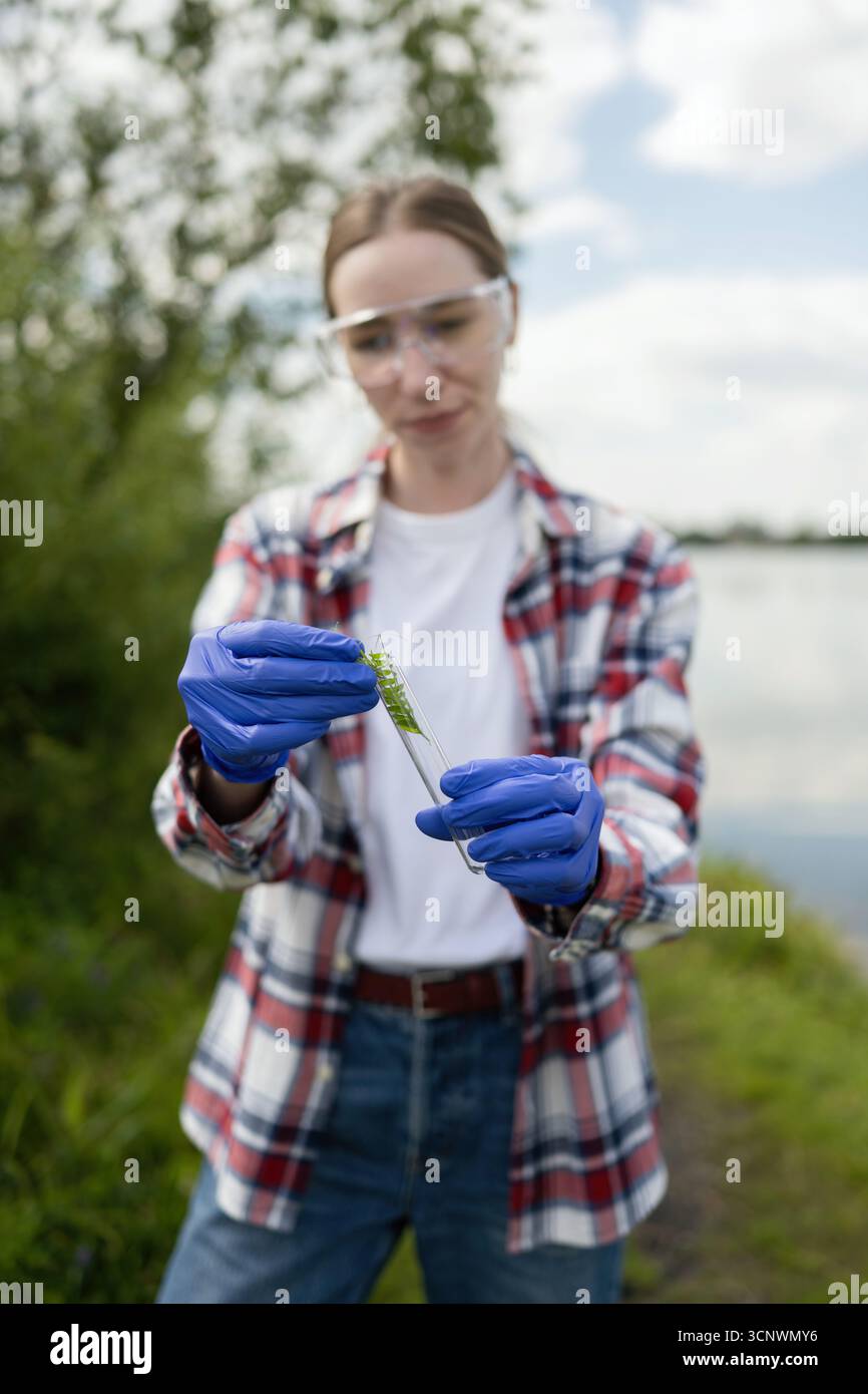 Una giovane biologa che indossa guanti e occhiali di protezione esamina un campione di piante in una provetta all'aperto, evidenziando la sua dedizione alla ricerca ecologica Foto Stock