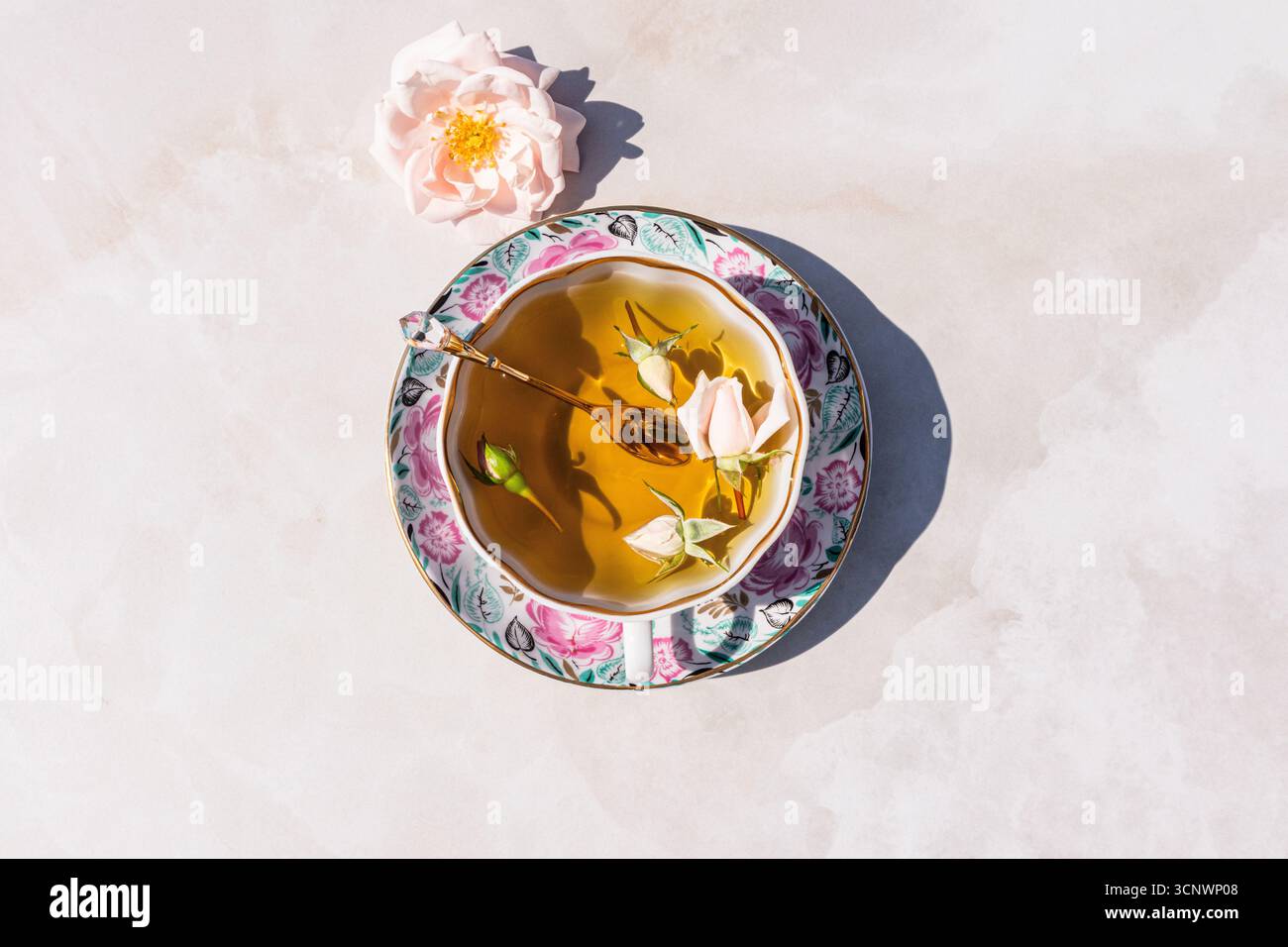 Una tazza di tè alle erbe splendidamente lavorata si trova elegantemente sopra un piattino dal design floreale, accompagnato da un delicato fiore rosa. La composizione trasuda caldo Foto Stock