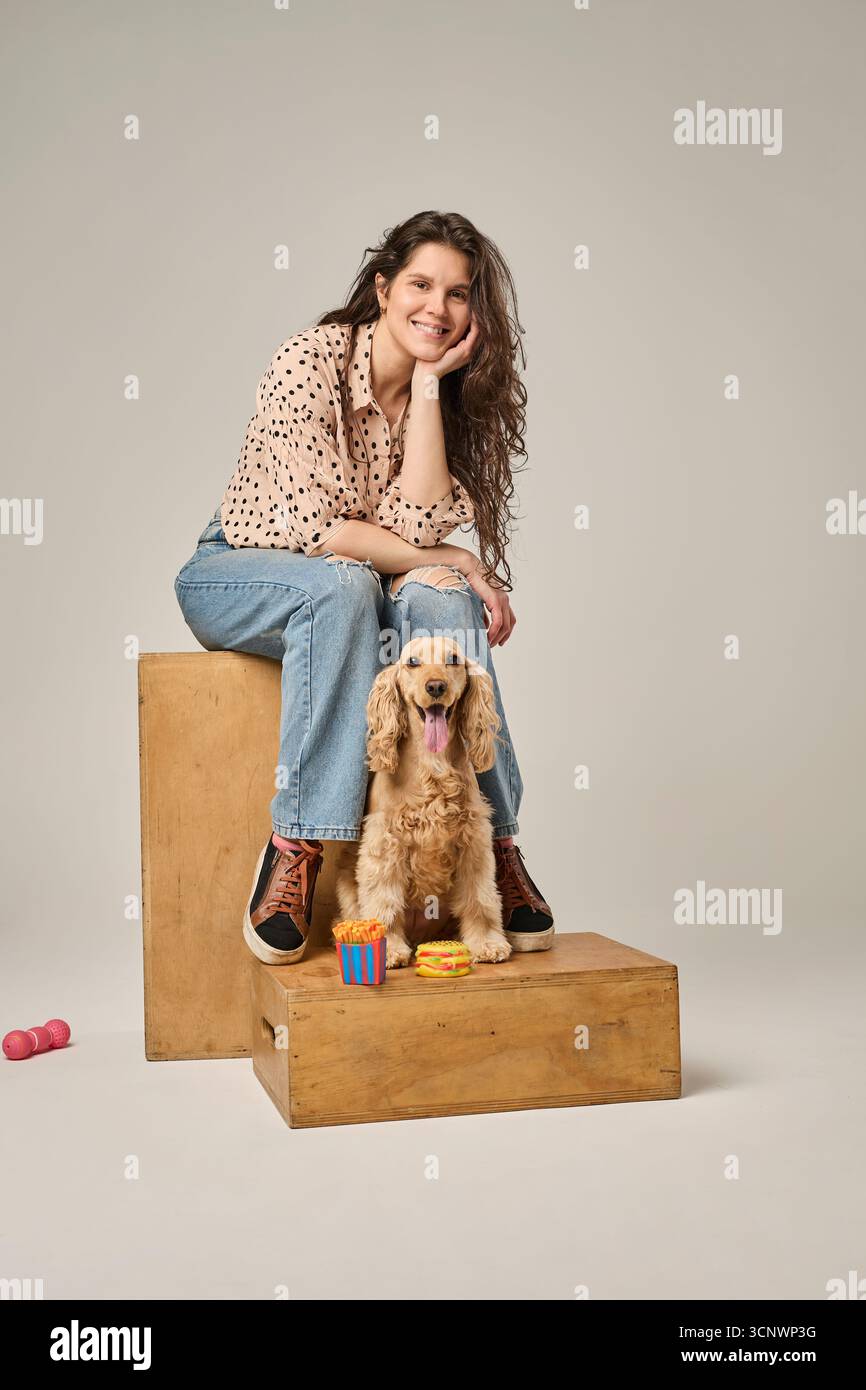 Una donna siede su una scatola di legno accanto a un felice cocker spaniel. Indossa jeans casual e una camicia a pois, creando un'atmosfera rilassata e amichevole, Foto Stock