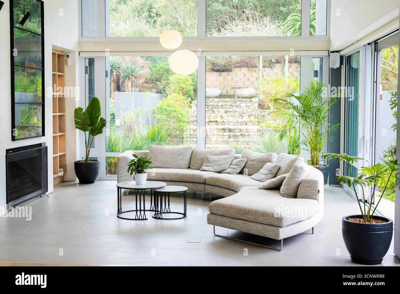 Divano sezionale curvo con porte in vetro, con tavolini da caffè con piante in vaso che si affacciano sul giardino Foto Stock