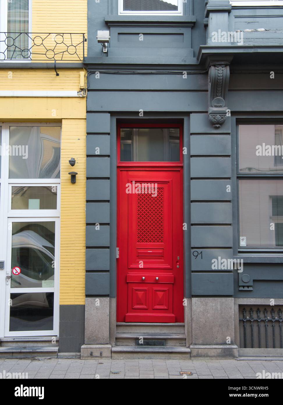 Vibrant Red Door su una facciata grigia - Una porta rossa brillante e audace con un motivo geometrico e delle maniglie delle portiere lucide si distingue contro una faca grigia a contrasto Foto Stock