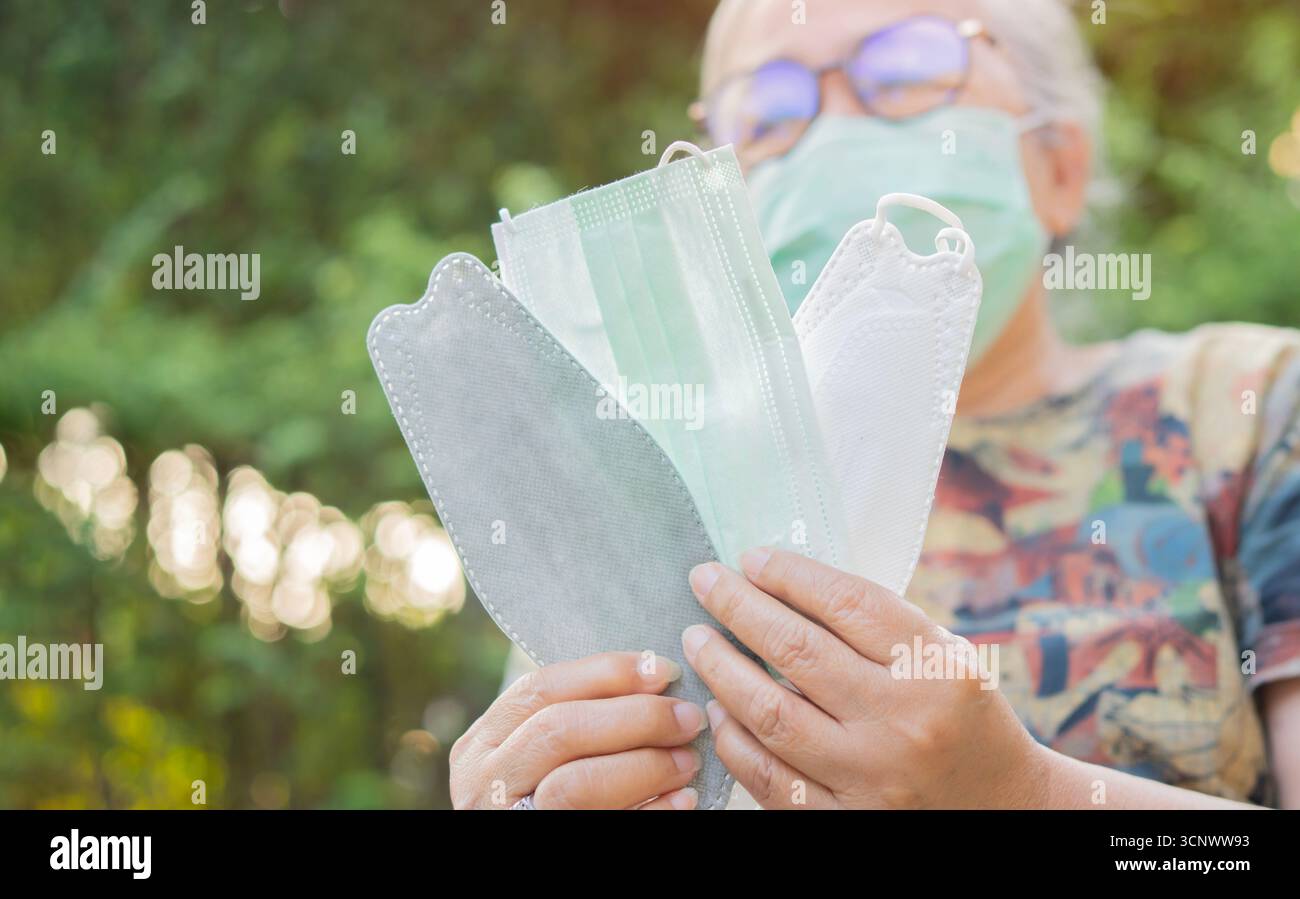 Donna anziana asiatica che tiene molte maschere mediche protettive sullo sfondo della natura. Medicina, cosmetologia e sicurezza Foto Stock