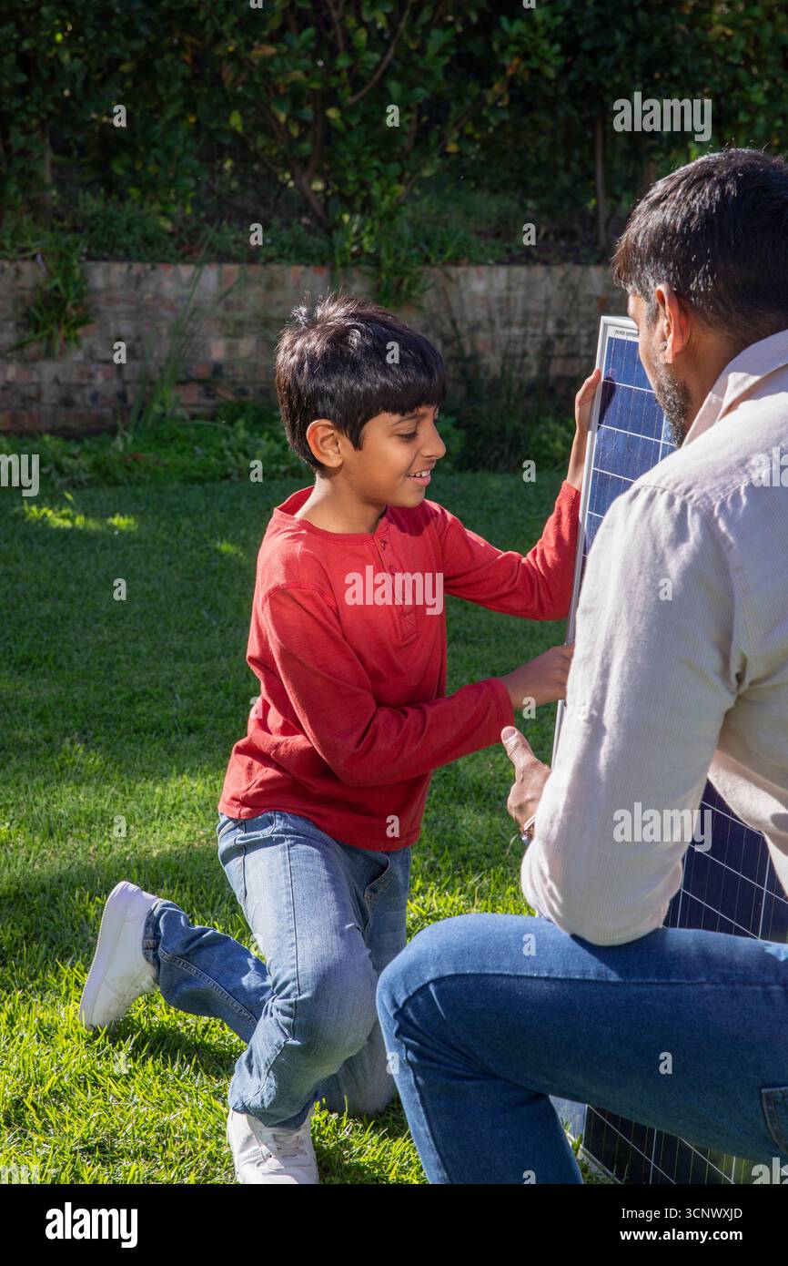 Padre asiatico con figlio inginocchiato che esamina il pannello solare nel cortile sotto il cielo soleggiato Foto Stock