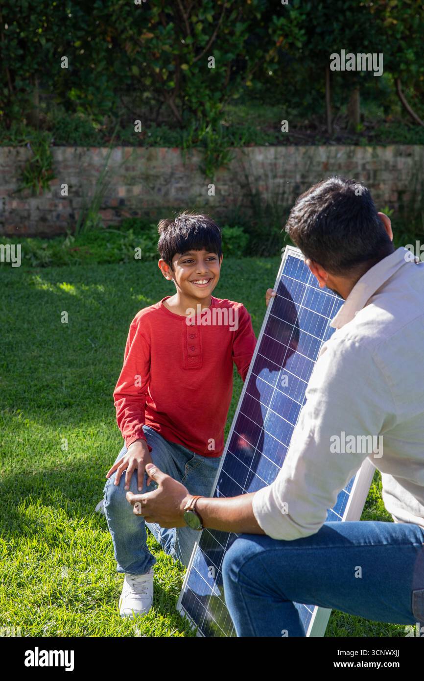 Padre e figlio indiani si inginocchiano sul prato del cortile con un grande pannello solare mentre imparano insieme Foto Stock