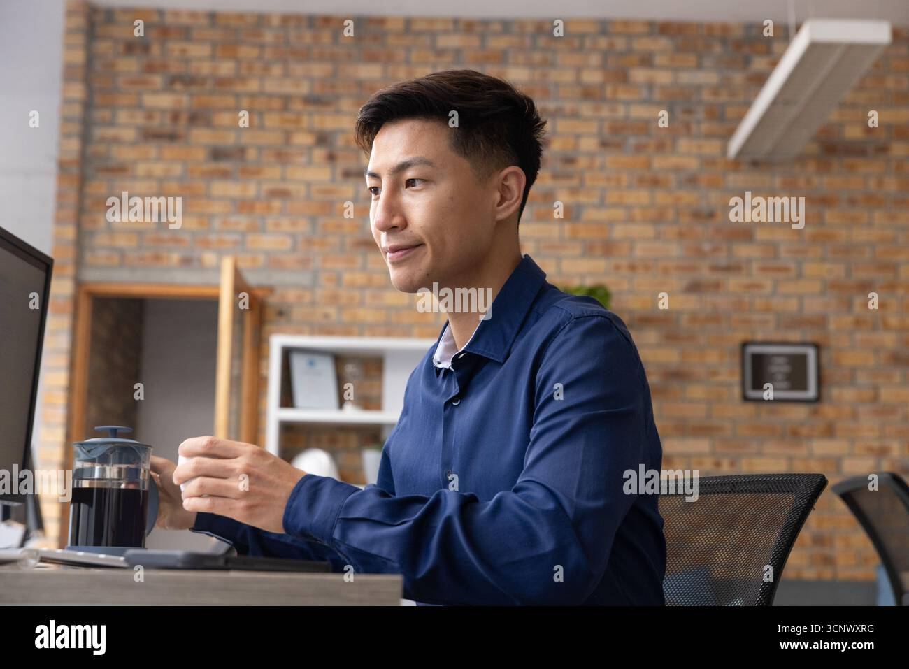 Uomo asiatico che prepara il caffè usando la stampa francese alla scrivania in ufficio contro il muro di mattoni, copia spazio Foto Stock