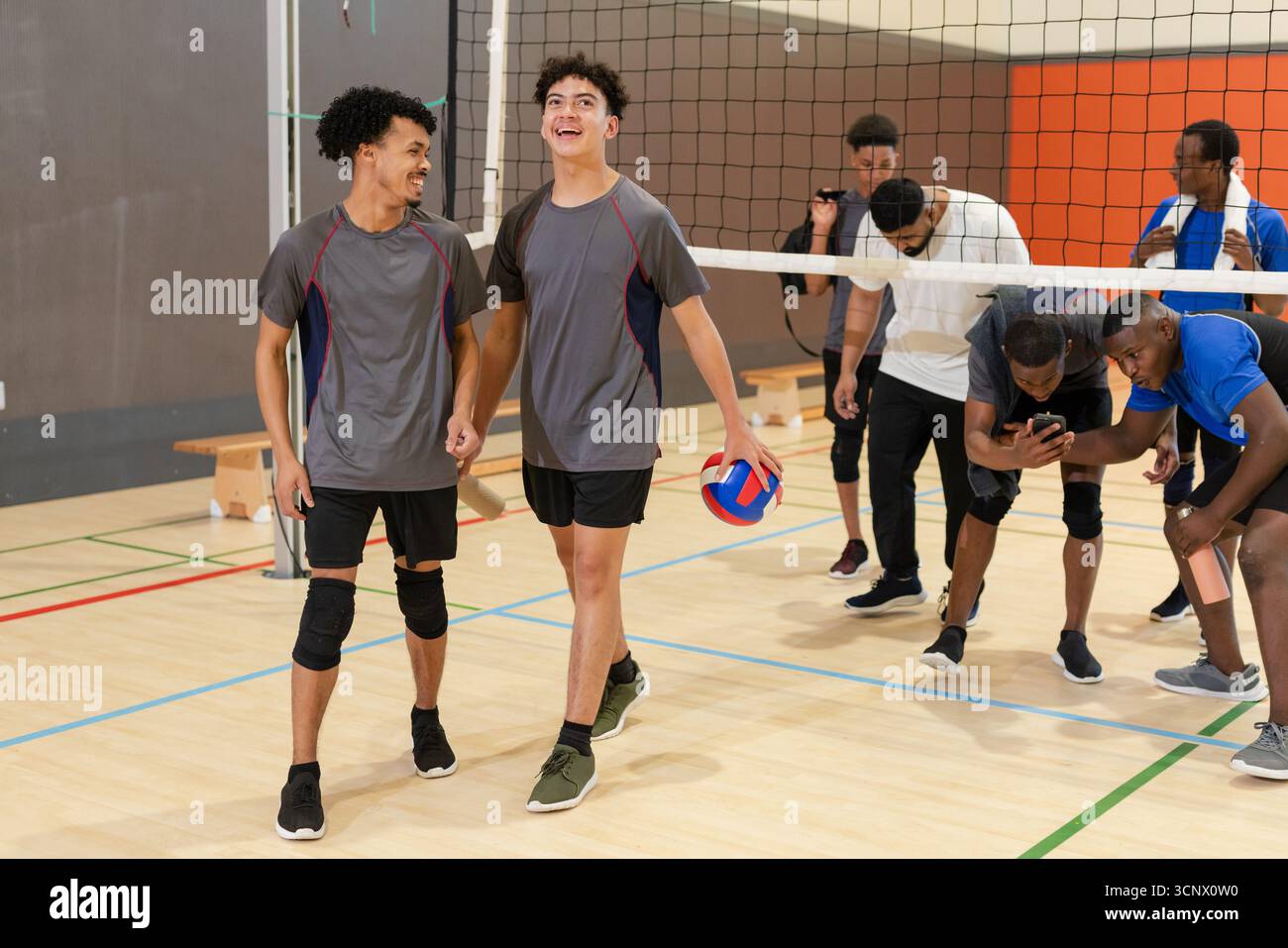 Compagni di squadra di pallavolo maschile che camminano in un abbigliamento sportivo sul campo tenendo la pallavolo rosso-bianco-blu accanto alla n Foto Stock