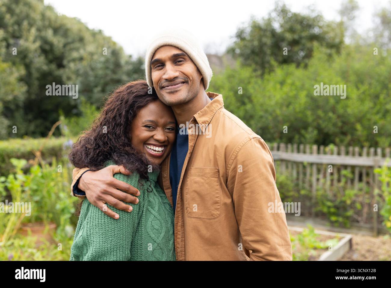 Coppia variegata che indossa un berretto in maglia e un pullover in maglia a cavo che si avvolgono nel cortile dietro la recinzione del picchetto Foto Stock