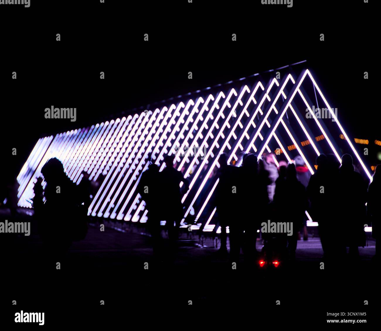 Le figure sagomate camminano davanti a un corridoio luminoso triangolare di notte, mescolando il movimento umano con l'illuminazione geometrica Foto Stock