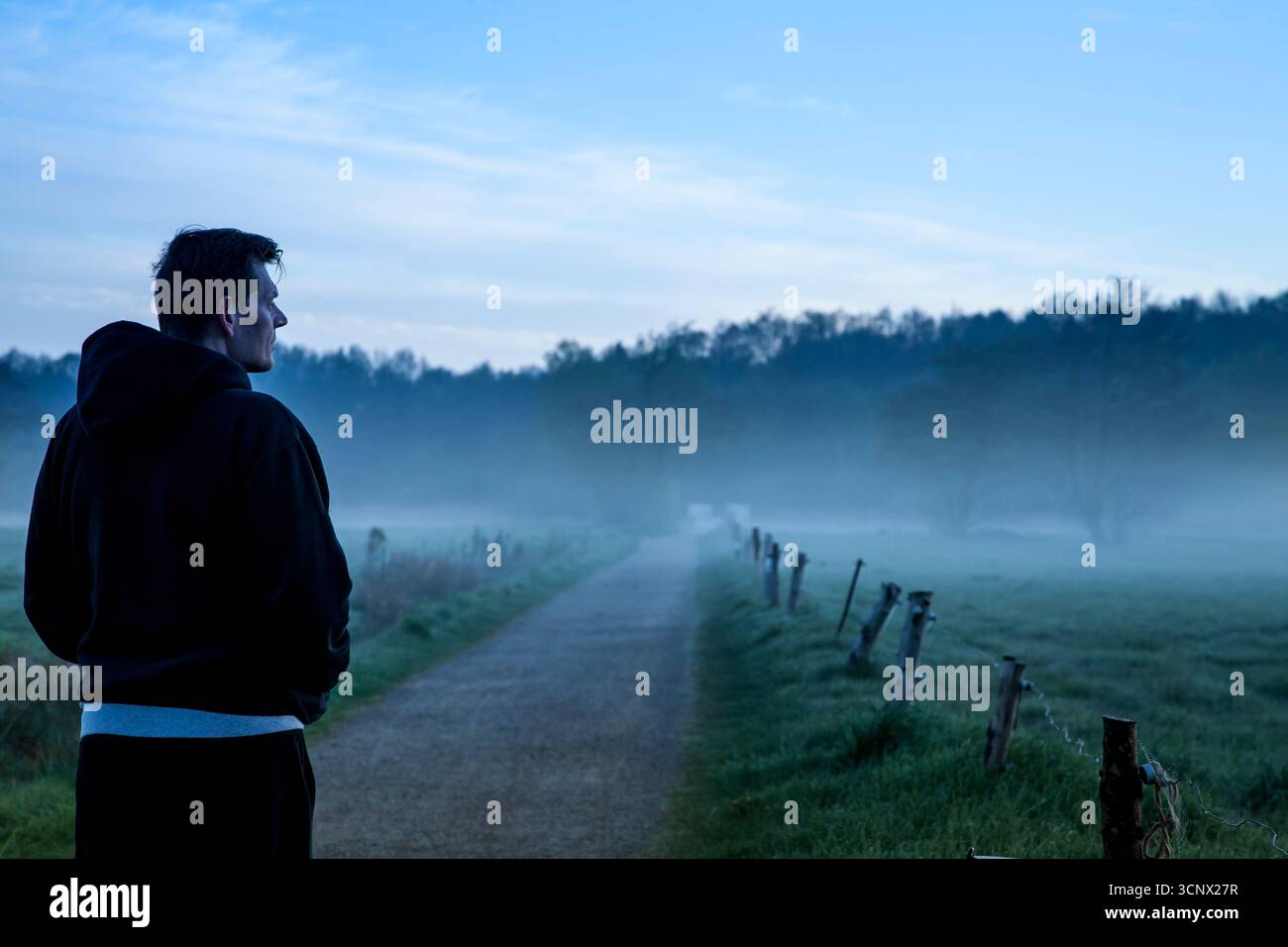 giovane uomo con back-to-camera in abbigliamento casual che guarda il paesaggio al mattino presto con nebbia e alba, uomo che pensa nella natura Foto Stock