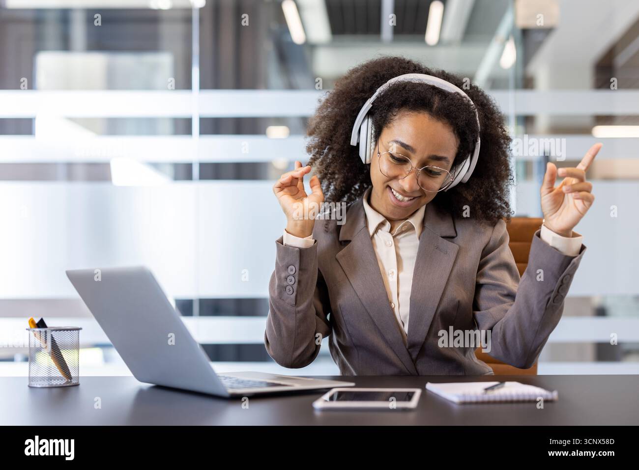 Giovane donna afroamericana, felice e rilassata, seduta alla scrivania in ufficio, con le cuffie, che ascolta la musica, balla seduto sulla sedia. Foto Stock