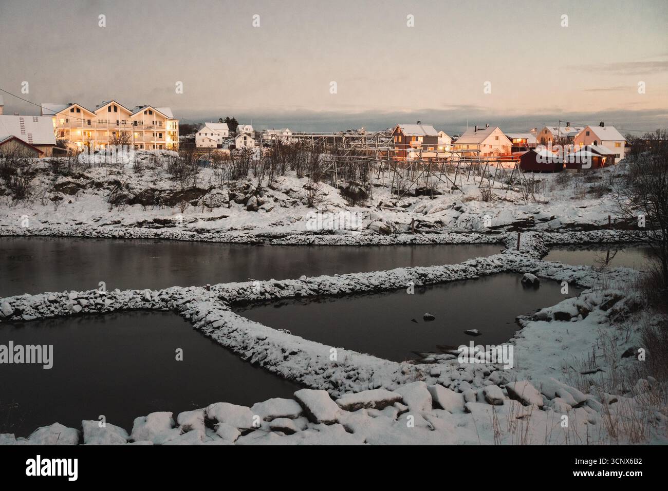 Idilliaco scenario invernale nelle isole Lofoten, Norvegia, con incantevoli case innevate che riflettono la luce dorata dell'inverno vicino a un tranquillo corso d'acqua, CA Foto Stock