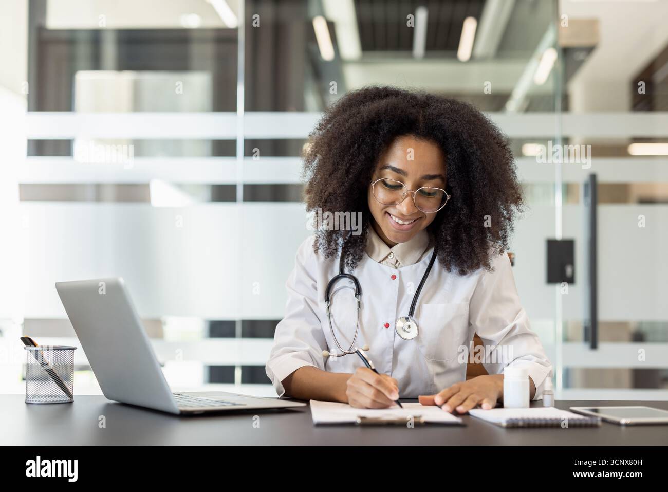 Una giovane dottoressa afroamericana è seduta a una scrivania in un abito e lavora su un computer portatile e con i documenti. Foto Stock