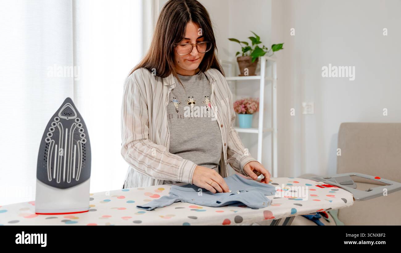Una donna incinta stirerà con cura i vestiti del bambino, preparandosi per l'arrivo del suo futuro bambino. Il suo tocco delicato e la sua concentrazione mostrano amore e anticipazione per lei Foto Stock