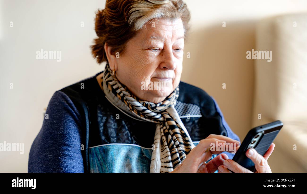 Una donna anziana siede in casa utilizzando uno smartphone, concentrandosi mentre naviga sullo schermo. L'immagine riflette l'uso della tecnologia, l'indipendenza e l'acti Foto Stock