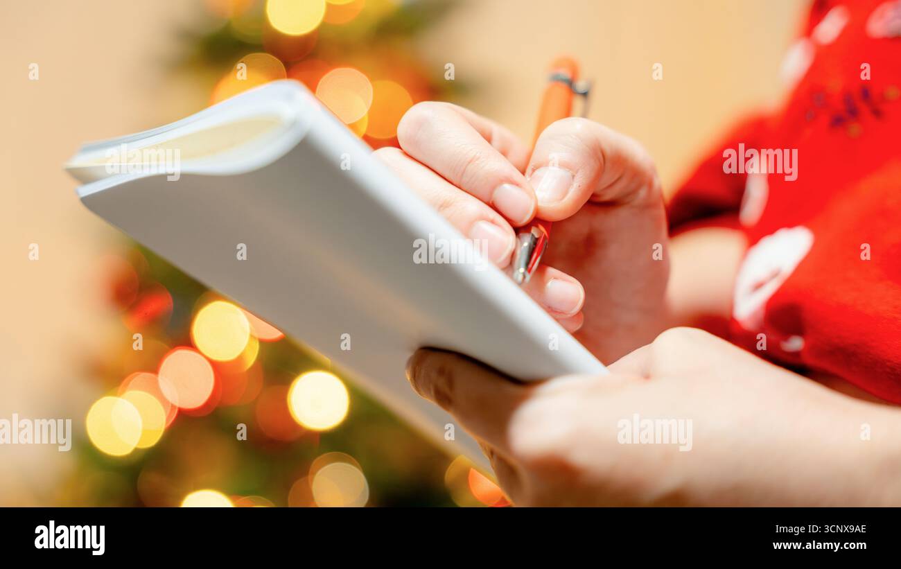 Primo piano ritagliato irriconoscibile delle mani di una persona che scrive su un taccuino, con un caldo albero di Natale sullo sfondo, per catturare un'atmosfera accogliente Foto Stock