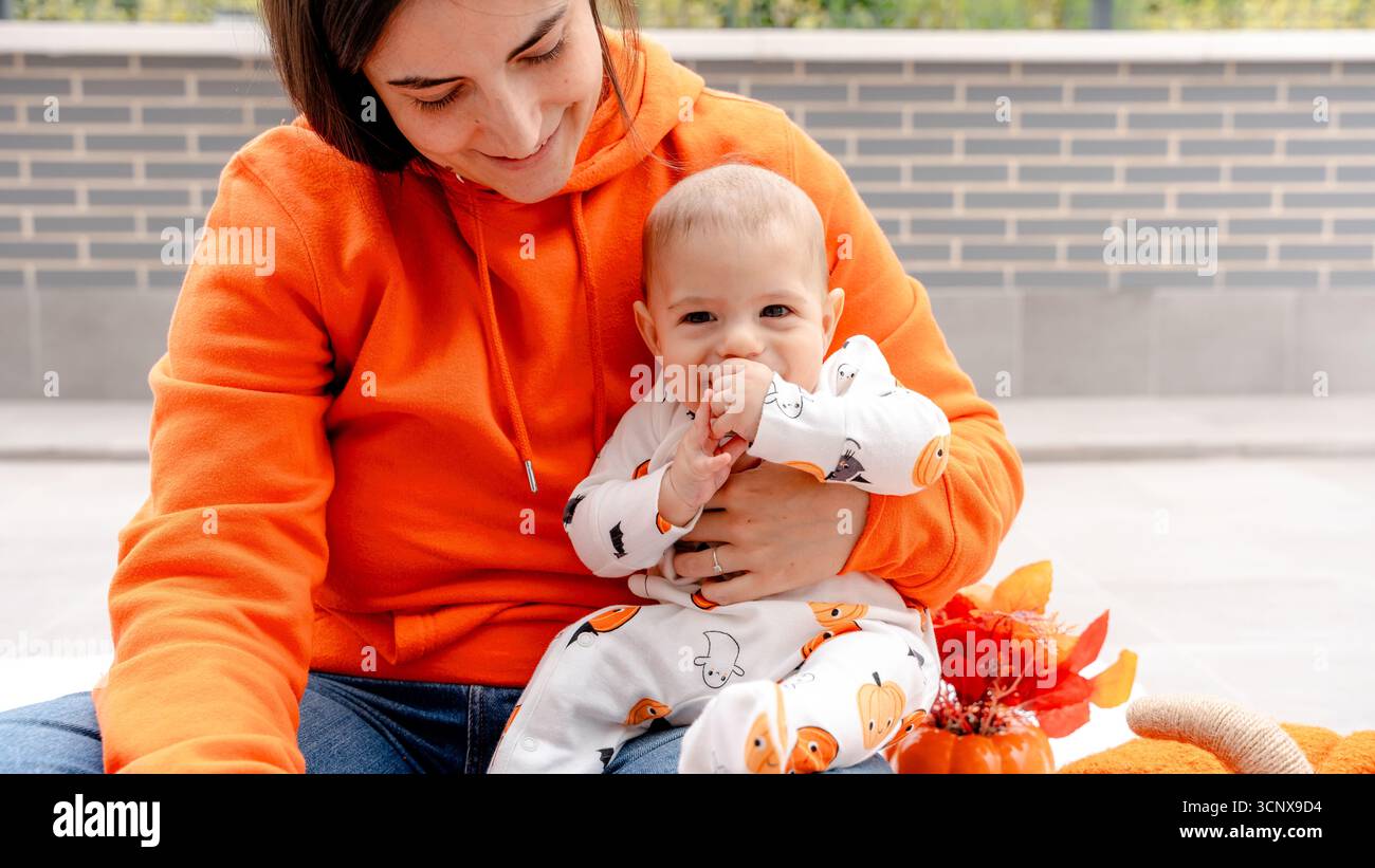 Una madre e il suo bambino di 4 mesi, vestiti con abiti a tema Halloween, godetevi un momento accogliente sulla terrazza. La madre abbraccia il suo bambino amorevolmente Foto Stock