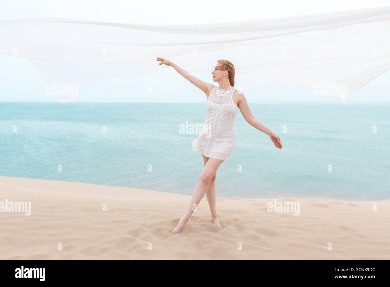 Elegante giovane donna in un abito bianco che scorre danzando graziosamente su una spiaggia di sabbia. Il tessuto si muove con il vento, incarnando la libertà e la bellezza dell'estate Foto Stock