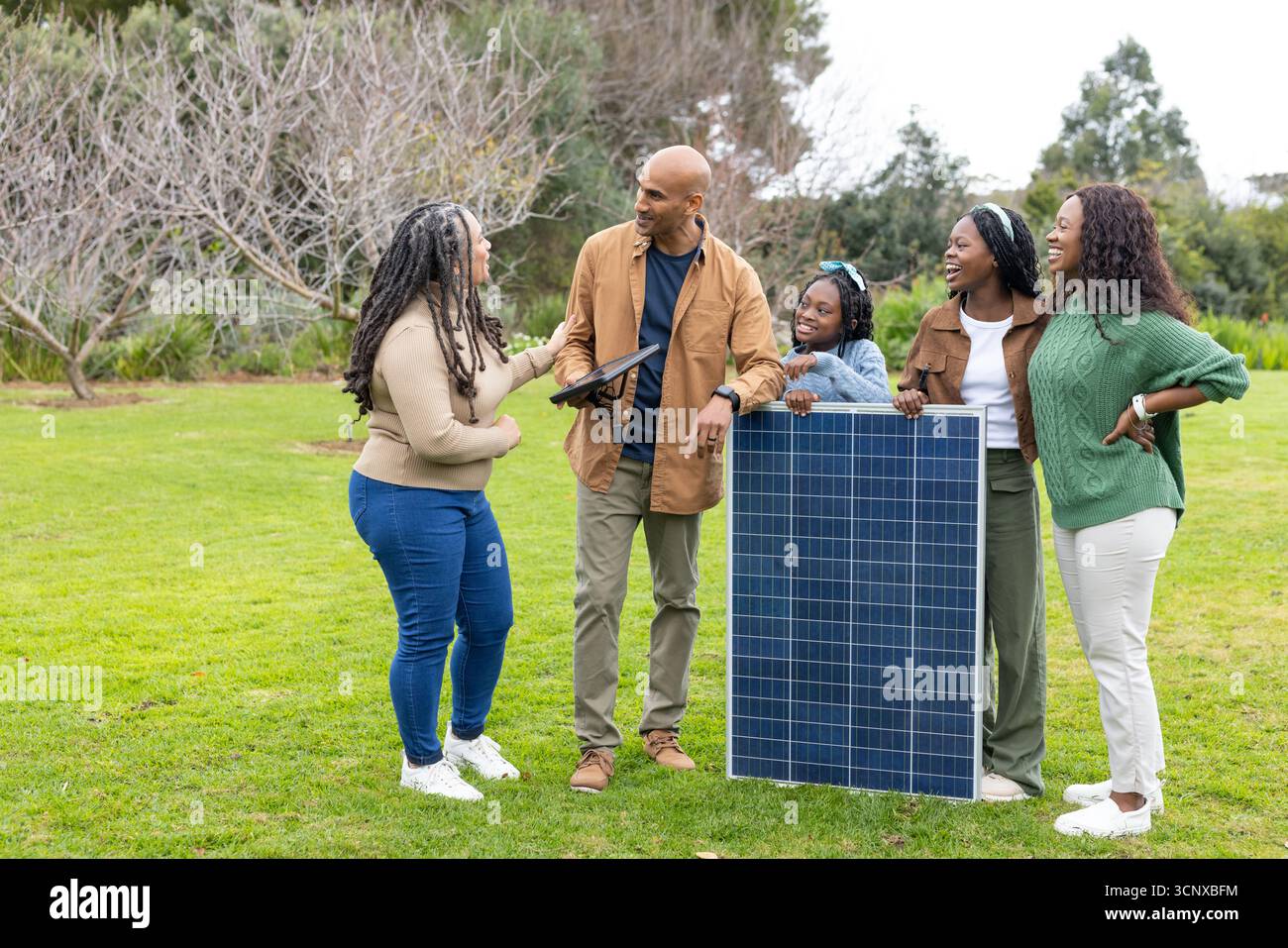 Famiglia diversificata che parla di pannelli solari su prati residenziali tenendo in mano un tablet touchscreen Foto Stock