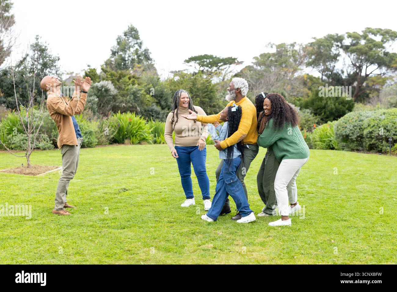 Famiglia diversificata di tre generazioni che lancia una piccola palla di gomma sul prato del cortile e sorride Foto Stock