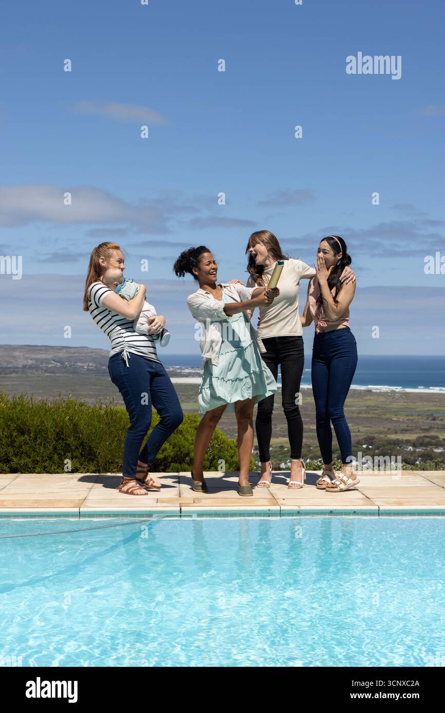 Diverse amiche che ridono, tengono in mano la bambola, il sonogramma sul ponte della piscina accanto alla piscina blu Foto Stock