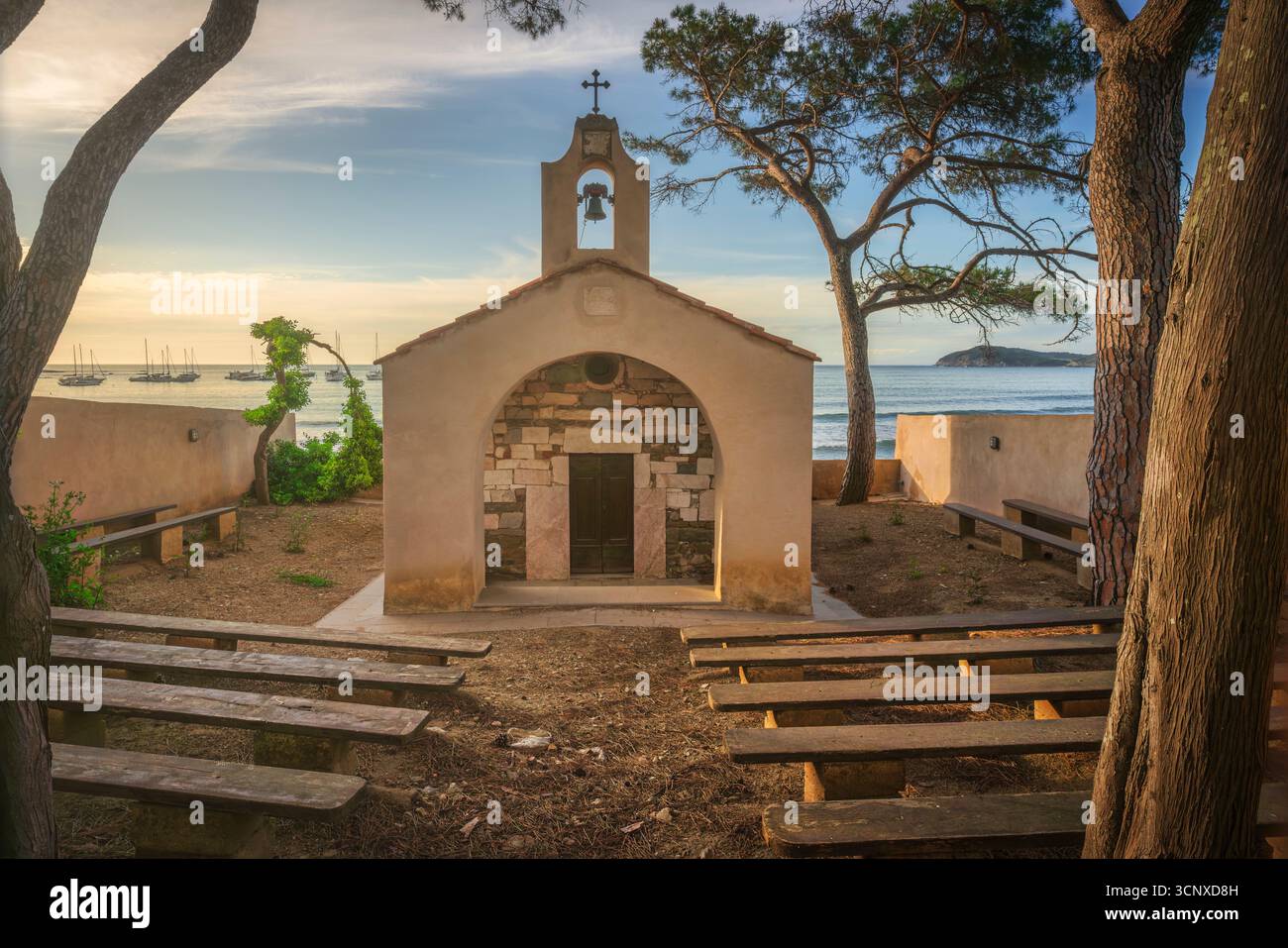 Storica cappella di San Cerbone che si affaccia sulla spiaggia di Baratti con barche a vela e il mare Mediterraneo. Piccola chiesa costiera incorniciata da pini in Toscana regio Foto Stock