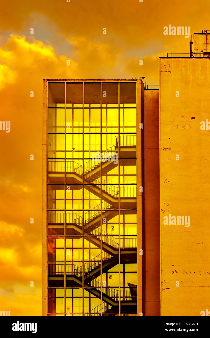Alte scale racchiuse in vetro con un suggestivo cielo arancione-giallo che si riflette sulla facciata durante il tramonto dell'università di Manchester nel Regno Unito Foto Stock
