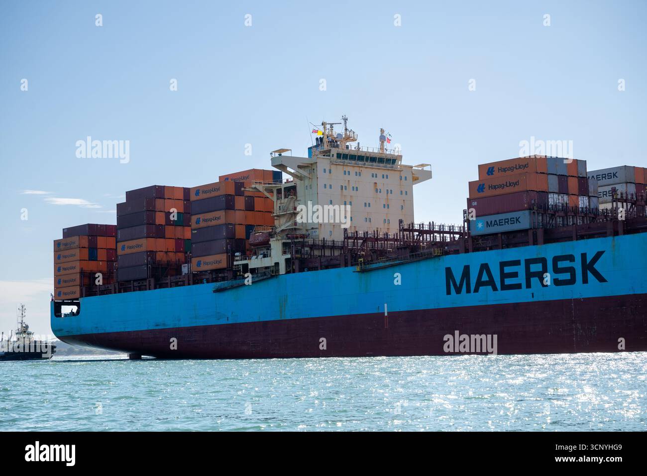 Una grande nave container di Maersk che trasporta numerosi container Hapag-Lloyd e Maersk naviga attraverso il mare, accompagnata da un piccolo rimorchiatore. Foto Stock