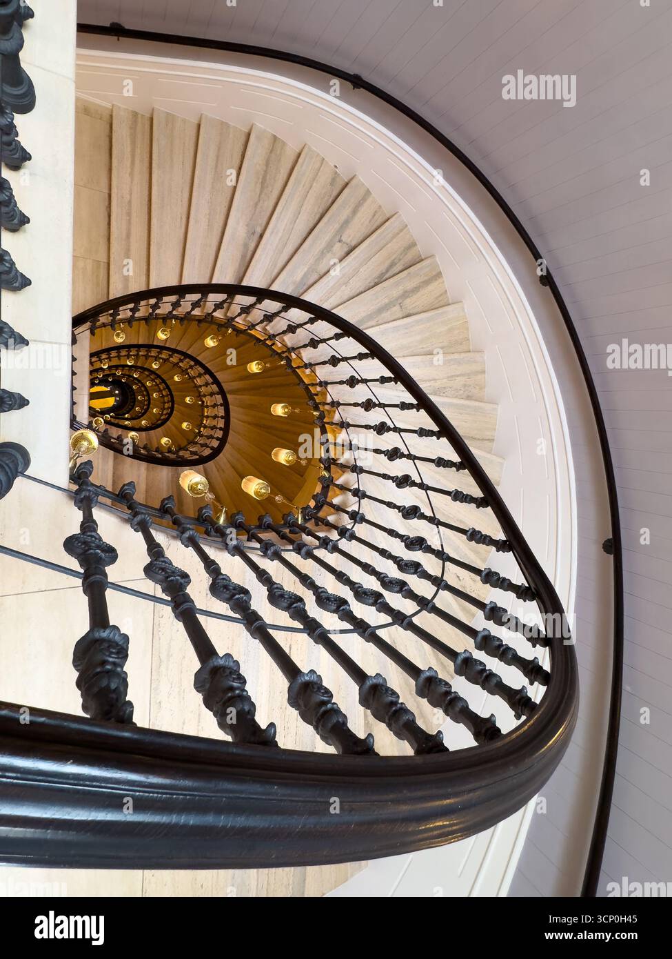 La scalinata a spirale in marmo vista dall'alto presenta balaustra nera ornata calda luce a lampadina che crea un ambiente interno classico ed elegante con profondità e movimento Foto Stock