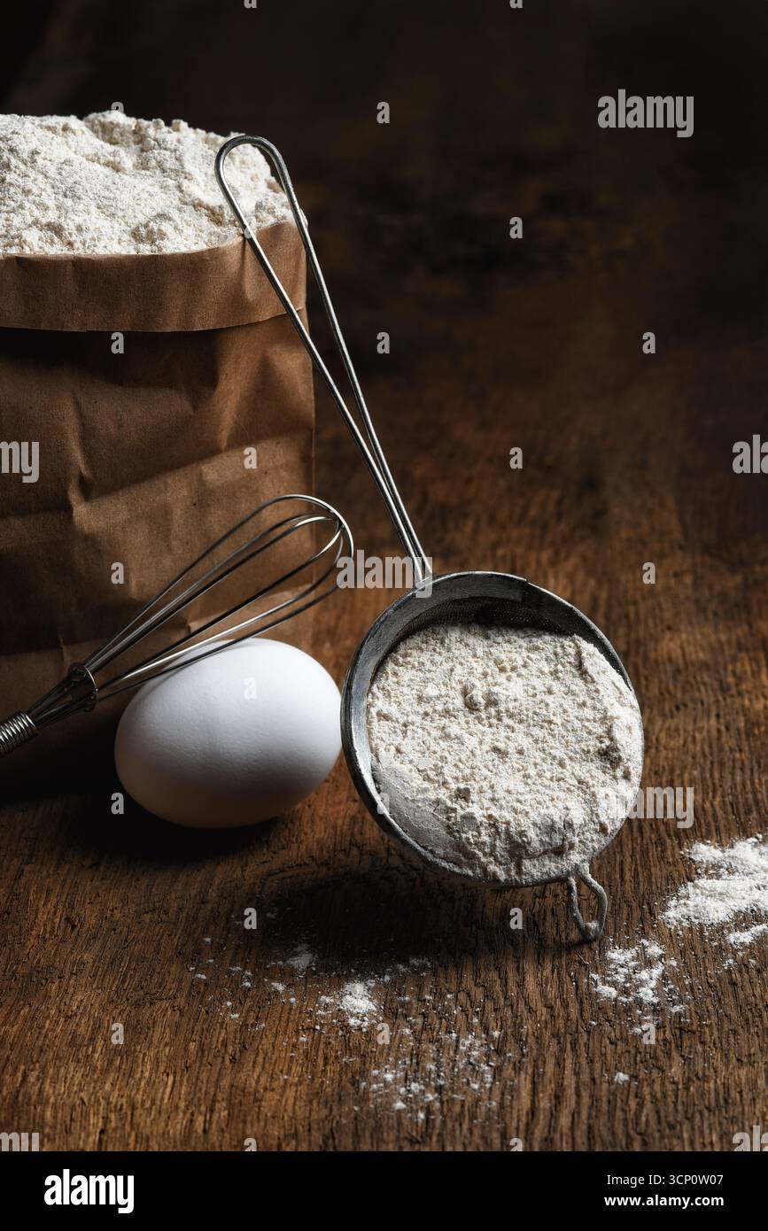 Cuocere la natura morta. Un sacchetto di farina con un setaccio pieno di farina appoggiato sul sacchetto, un uovo e una frusta hanno scoperto la scena su un rustico tavolo di legno. Foto Stock