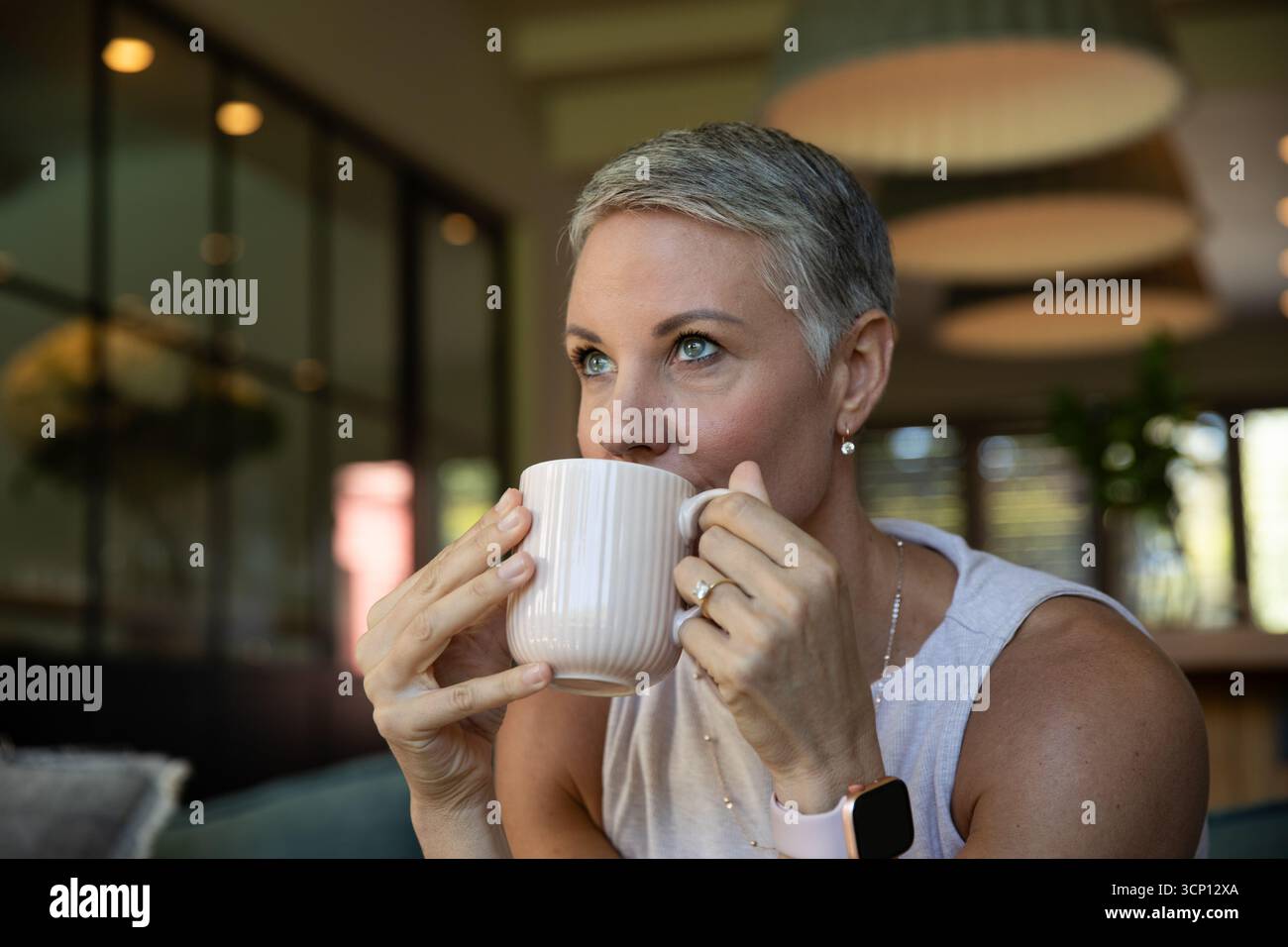 Donna di mezza età che sorseggia una tazza a coste mentre indossa lo smartwatch e la collana al tavolo da caffè Foto Stock