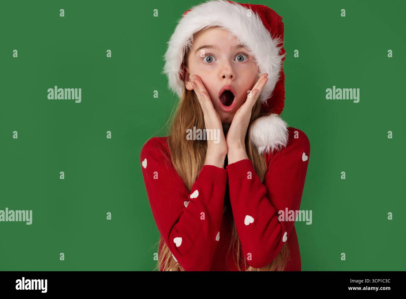 Una ragazza sorpresa con un cappello di Babbo Natale e un maglione rosso tiene la faccia sotto shock su uno sfondo verde Foto Stock