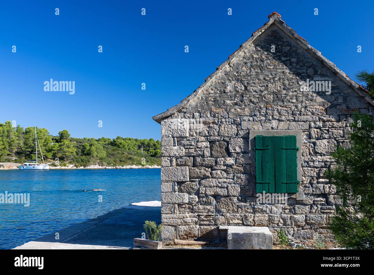 Un rustico cottage in pietra si trova sul bordo dell'acqua con vivaci persiane verdi. Le calme acque blu, gli alberi di pino e le barche distanti creano una costa tranquilla Foto Stock