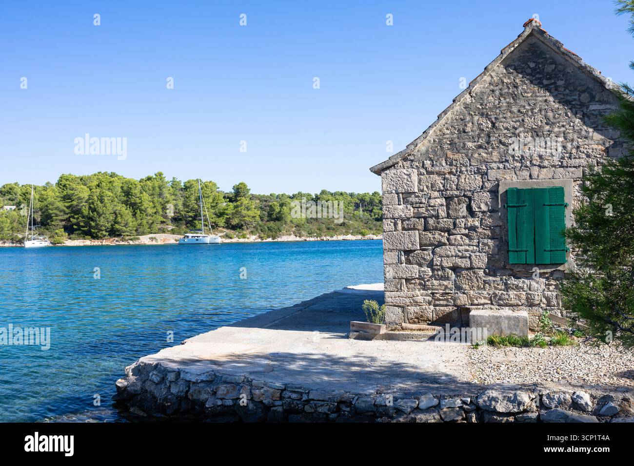 Un rustico cottage in pietra si trova sul bordo dell'acqua con vivaci persiane verdi. Le calme acque blu, gli alberi di pino e le barche distanti creano una costa tranquilla Foto Stock