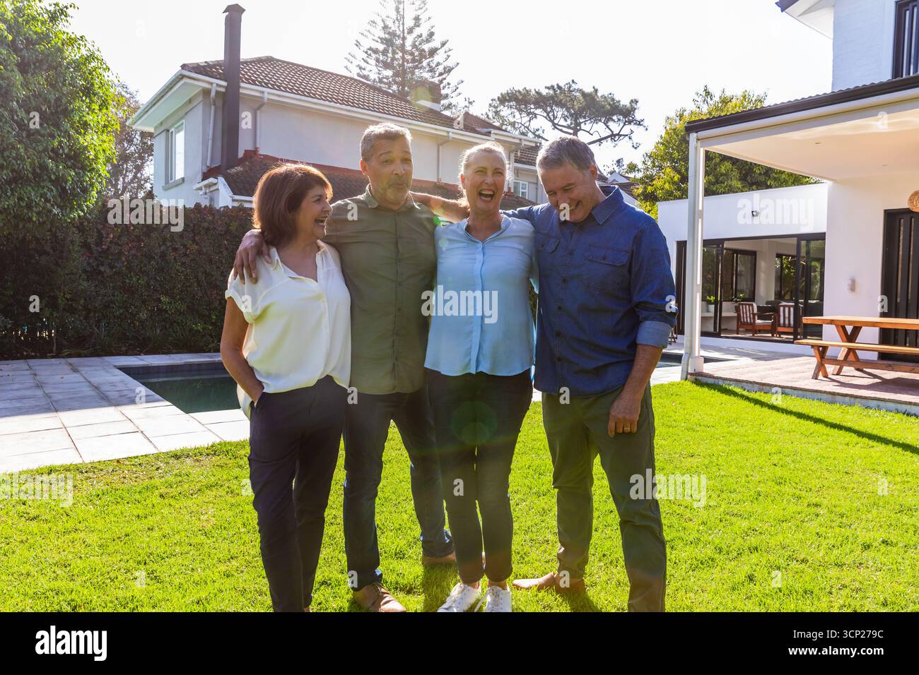 Amici di mezza età che ridono mentre si trovano sul prato verde accanto alla terrazza con piscina e al patio piastrellati Foto Stock