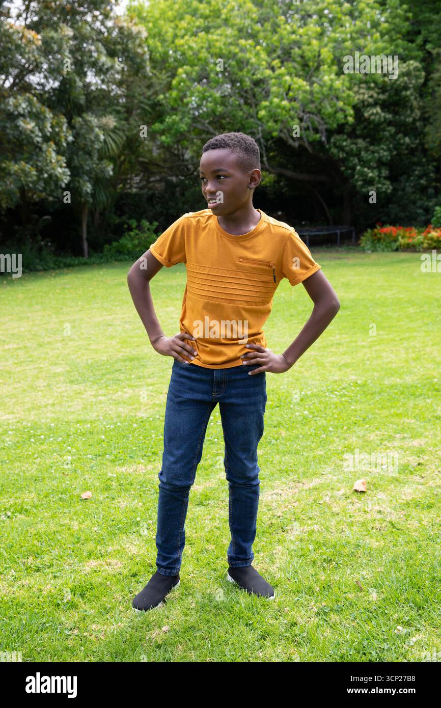 Ragazzo afroamericano in piedi sul prato accanto alla panchina di legno al parco con t-shirt e jeans Foto Stock