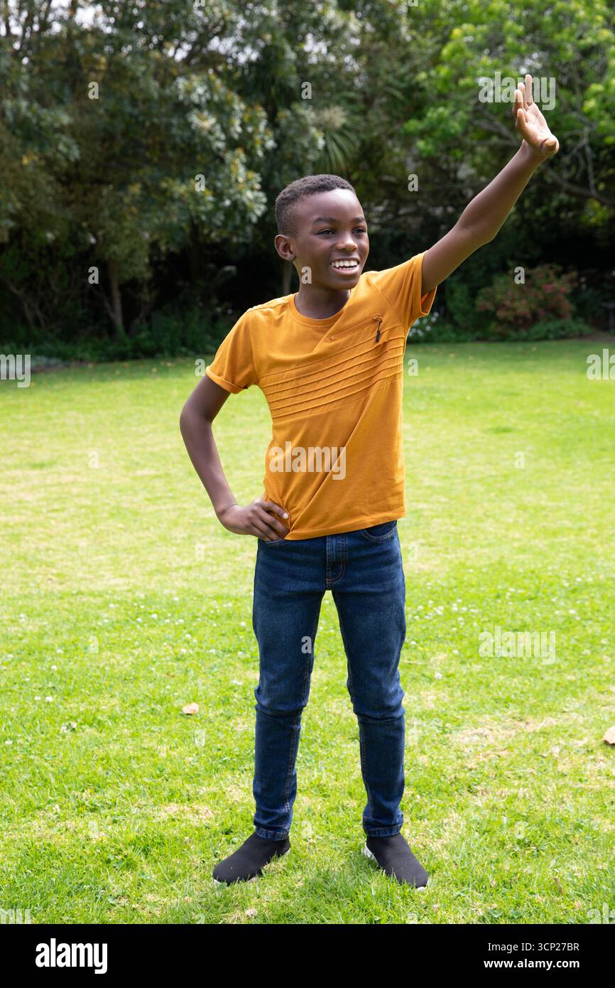 Ragazzo afroamericano che sventola, posa con la mano sull'anca in giardino indossando una t-shirt arancione, jeans Foto Stock