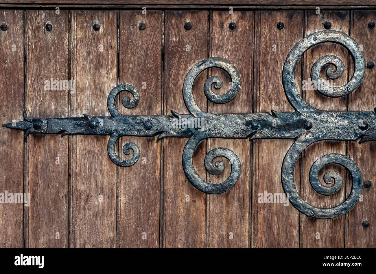 Dettaglio di cerniera in ferro battuto ornato sulla porta storica della chiesa, catturato da una luce dorata soffusa con una rustica struttura in legno. Foto Stock
