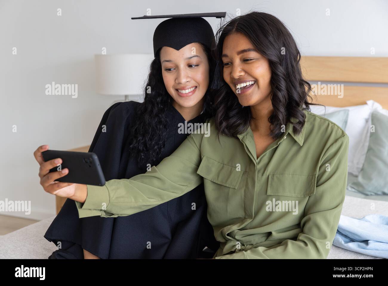 Mamma e figlia adolescente diversificate che scattano selfie in camera da letto con cappello, abito e smartphone Foto Stock