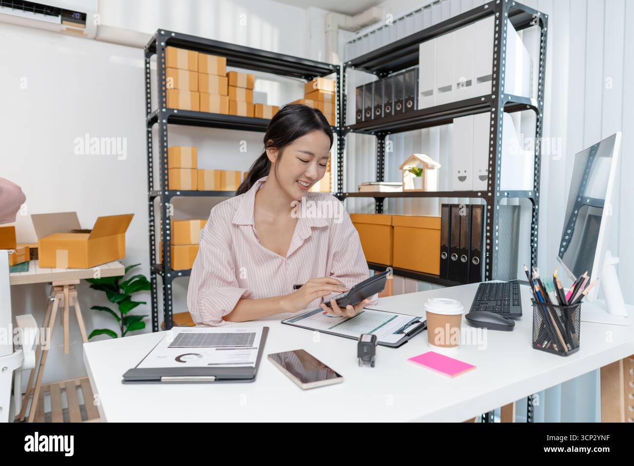 Una giovane donna con una camicia a righe lavora alla sua scrivania con documenti, calcolatrice e smartphone, concentrandosi sulle attività contabili e sulla pianificazione finanziaria Foto Stock