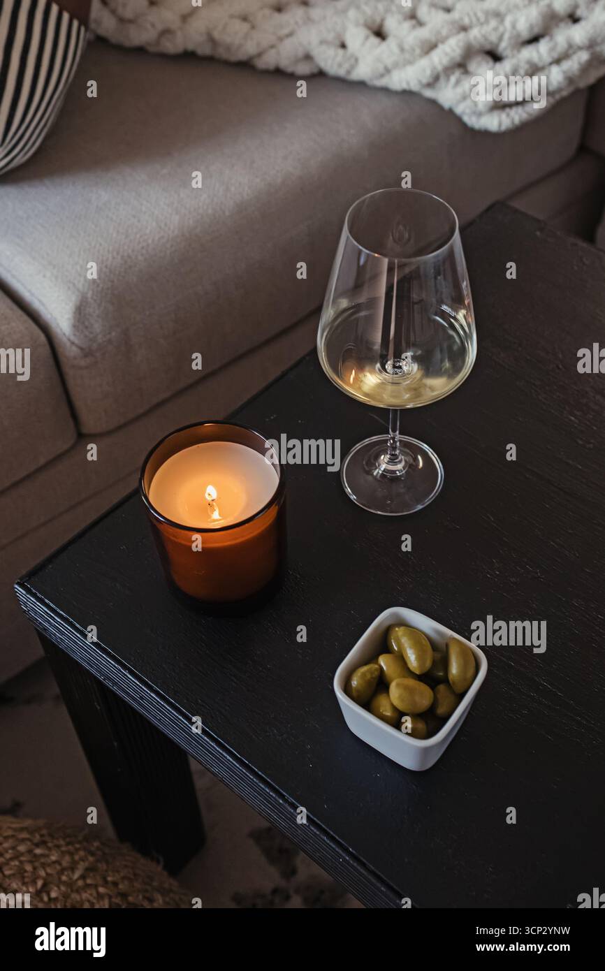 Una scena rilassante con un bicchiere di vino bianco, una candela illuminata e una ciotola di olive su un tavolo di legno scuro. Foto Stock