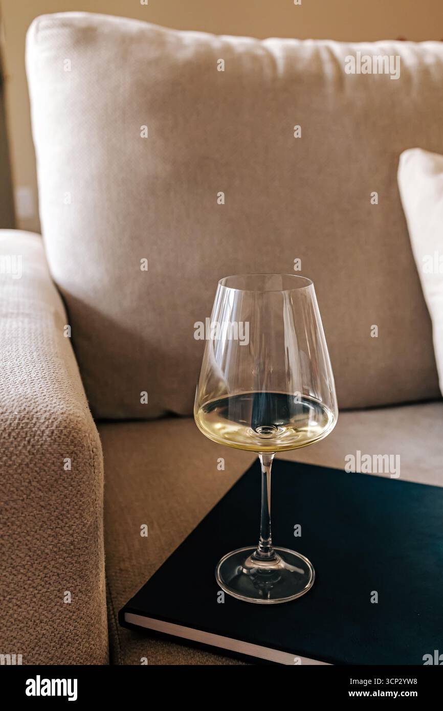 Un bicchiere di vino bianco riposa su un libro chiuso in cima a un morbido divano beige, creando un'atmosfera accogliente e rilassante perfetta per rilassarsi a casa. Foto Stock