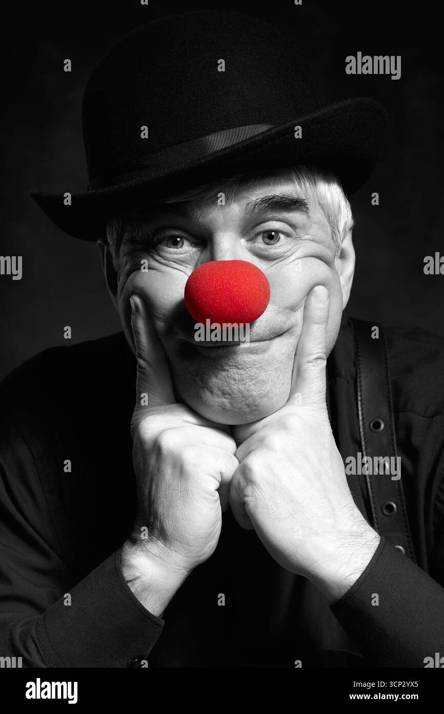 Spettacolare ritratto a colori selettivo di un pagliaccio triste su sfondo scuro. Un uomo anziano con naso rosso da clown e cappello da bowler sulla testa. Vestito con blac Foto Stock