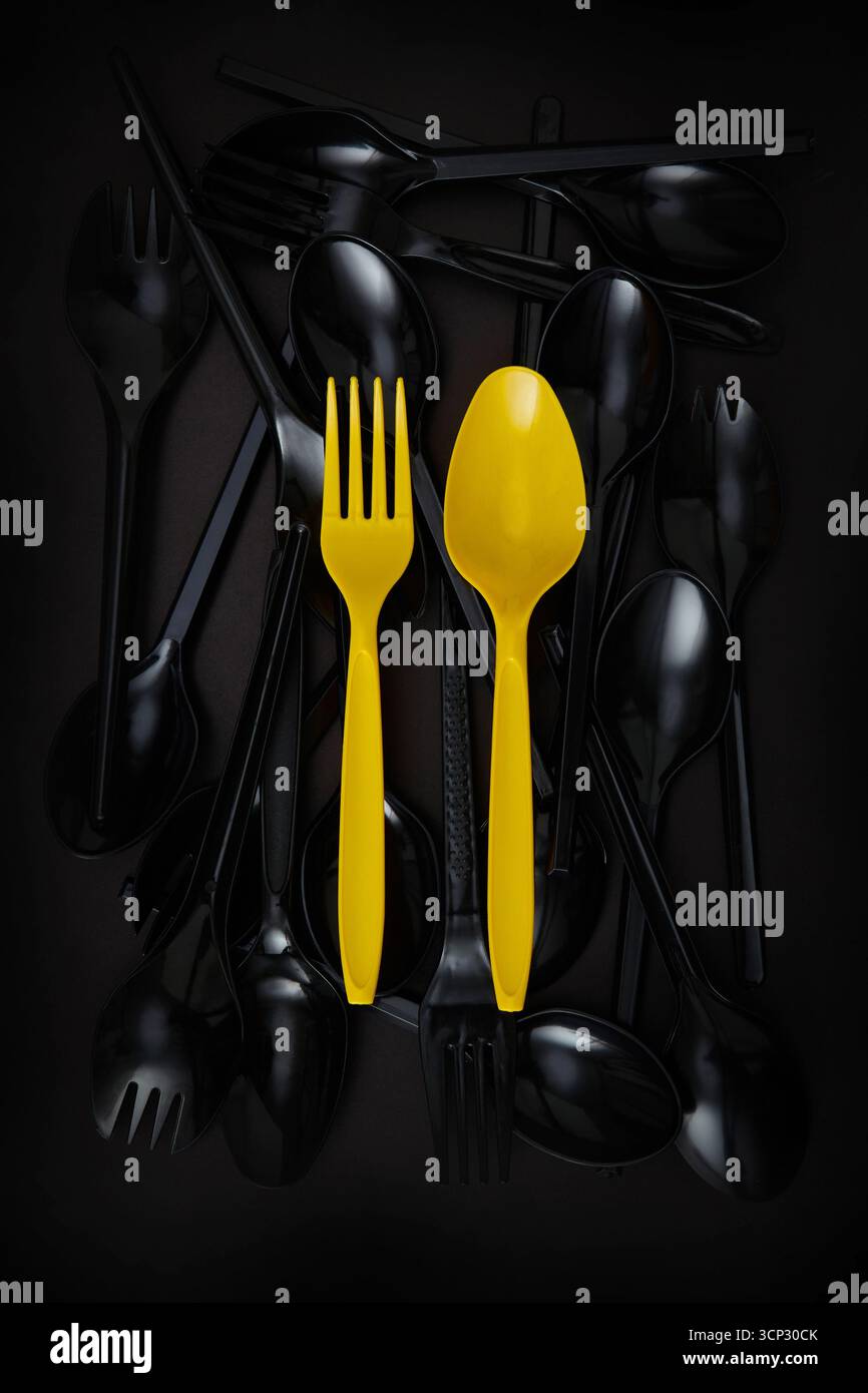 Una collezione di cucchiai e forchette di plastica nera con due utensili gialli luminosi, una forchetta e un cucchiaio, che si distinguono in modo prominente con sfondo scuro Foto Stock