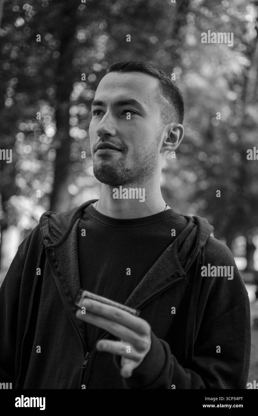 Un giovane sta suonando un'armonica mentre sta in piedi su un sentiero in un parco verde. Alberi alti forniscono uno sfondo naturale mentre la luce del sole filtra attraverso il lea Foto Stock