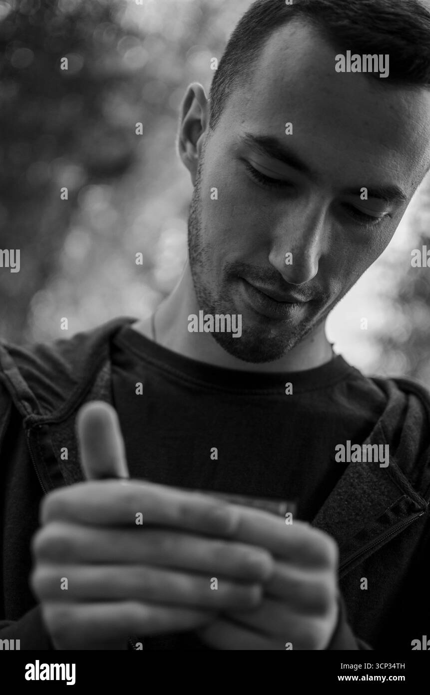 Un giovane sta suonando un'armonica mentre sta in piedi su un sentiero in un parco verde. Alberi alti forniscono uno sfondo naturale mentre la luce del sole filtra attraverso il lea Foto Stock