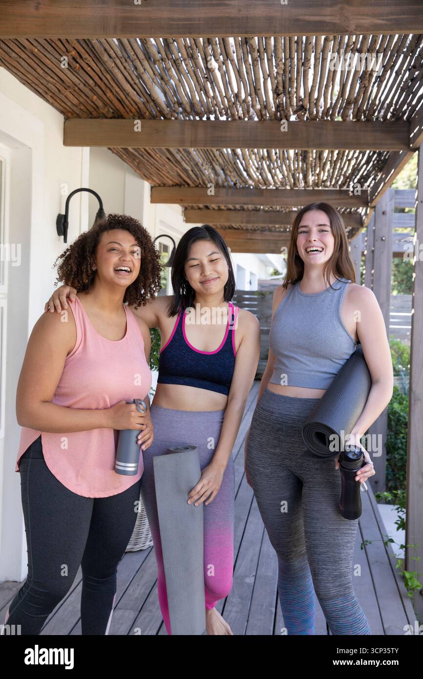 Diverse amiche in abbigliamento sportivo sorridenti con stuoie da yoga e bottiglie d'acqua sotto il pergolato sul ponte Foto Stock